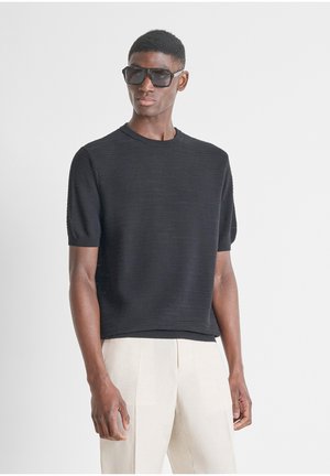 Man die een zwart, kort mouw gebreid shirt draagt, beige broek en grote zwarte zonnebril, staand voor een eenvoudige lichte achtergrond.