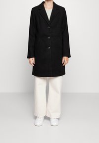 Cappotto in misto lana nero con rever a punta, cinque bottoni e tasche laterali, abbinato a pantaloni ampi beige chiaro e sneakers bianche.