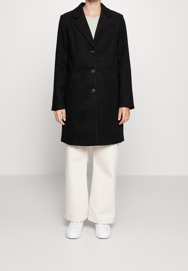 Cappotto in misto lana nero con rever a punta, cinque bottoni e tasche laterali, abbinato a pantaloni ampi beige chiaro e sneakers bianche.