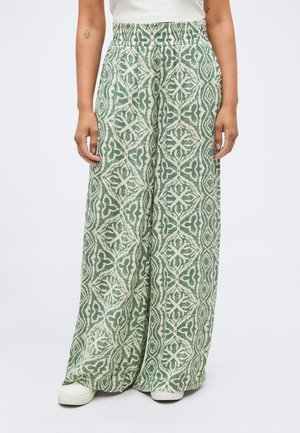 Παντελόνια με φαρδιά πιέτα, διακοσμημένα με πολύπλοκα γεωμετρικά σχέδια σε πράσινο και λευκό, φορεμένα με λευκά παπούτσια και λευκό μπλουζάκι.