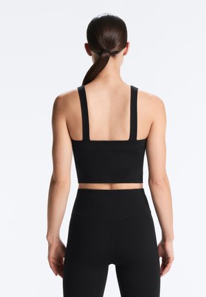 Frau mit dunklem Pferdeschwanz, die ein schwarzes, ärmelloses Crop-Top und hochtaillierte Leggings trägt, steht mit dem Rücken zum Betrachter vor einem schlichten weißen Hintergrund.