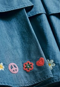 Denimstoffdisplay mit gestickten Patches: pinkes Friedenszeichen, rote Blume, rotes Herz und gelbe sowie weiße Gänseblümchen entlang des Saums.