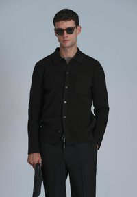 Schwarze Strickjacke mit Kragen, Knopfverschluss, einer Brusttasche und langen Ärmeln, kombiniert mit dunklen Hosen und hält einen schwarzen Gegenstand.