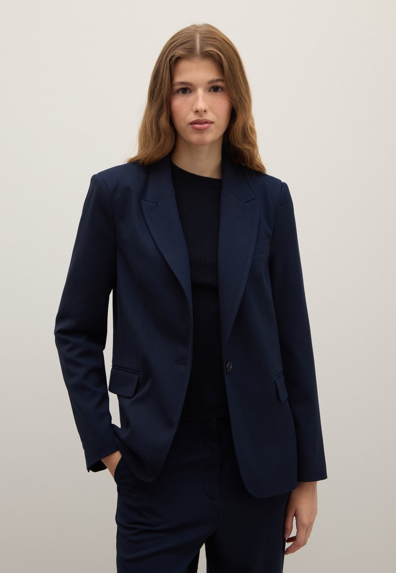 Blazer blu navy in tessuto liscio, con colletto a rever, chiusura con un solo bottone e due tasche frontali. Abbinato a una maglia nera a costine.