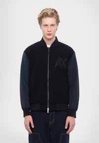 Giubbotto bomber nero con maniche blu navy, chiusura con zip, polsini a coste, tessuto strutturato, con logo AX in rilievo sulla parte anteriore.