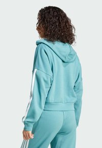 Turkoosi huppari, jossa on rento leikkaus, valkoiset sivuviivat ja ribattuja hihansuita. Pehmeä kangas, jossa on sileä pinta ja minimalistinen muotoilu.