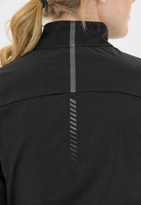 Svart sportjacka med hög krage och reflekterande vertikala detaljer på baksidan för synlighet och stil. Slät textur.