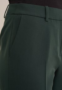 Tmavě zelené kalhoty na míru s hladkým povrchem, které mají přední kapsy a plochý design pasu. Detailní pohled zvýrazňuje strukturu látky.