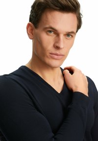 Navyblaues V-Ausschnitt-Pullover mit gerippter Textur und enganliegenden Ärmeln, getragen von einer Person mit kurzen Haaren, die ihre Hand auf die Schulter legt.