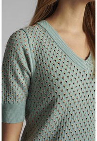 Pull à col en V vert clair avec des trous texturés et des accents corail. Manches courtes avec poignets côtelés. Tissu en maille lisse.