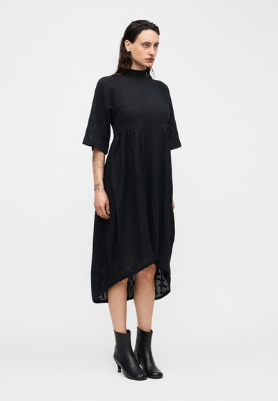 Γυναίκα που φοράει μαύρο midi φόρεμα με ψηλό λαιμό και μανίκια μέχρι τον αγκώνα, μαζί με μαύρα μποτάκια μέχρι τον αστράγαλο, στέκεται μπροστά σε απλό λευκό φόντο.