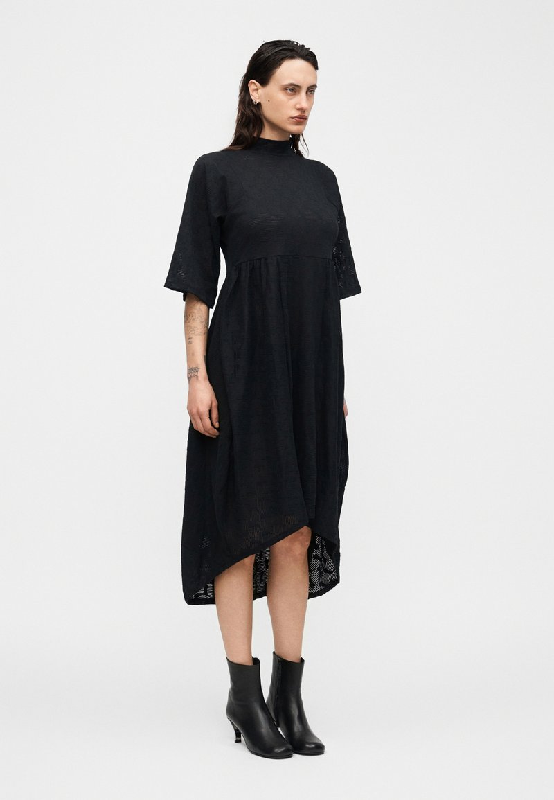 Frau trägt ein schwarzes Midikleid mit hohem Kragen, Ellbogenlangen Ärmeln und schwarzen Stiefeletten, steht vor einem schlichten weißen Hintergrund.