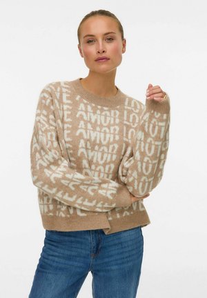 Vrouw met haar vastgebonden, gekleed in een beige trui met witte teksten "LOVE" en "AMOR" en blauwe spijkerbroek, staand tegen een witte achtergrond.