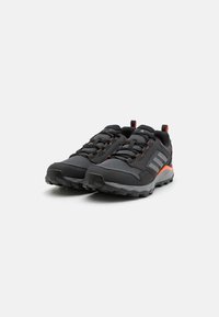 Baskets de sport noires avec un matériau en mesh, des accents orange et une semelle durable avec des crampons profonds pour une meilleure traction. Détails réfléchissants présents.