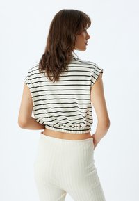 Korte, cropped top in crème met zwarte horizontale strepen, voorzien van een elastische band bij de taille. Gecombineerd met ribgebreide, lichtgekleurde broeken.