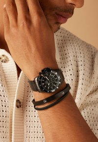 Schwarze Chronograph-Uhr mit metallischem Armband, weißen Akzenten und mehreren Zifferblättern. Zwei schwarze Armbänder: eines glatt, das andere geflochten mit einem Metallelement.