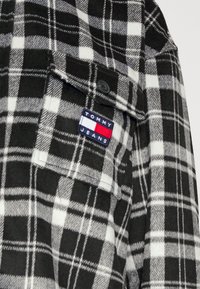 Black and white plaid flannel shirt with a chest pocket. Features a fabric patch labeled "TOMMY JEANS" in navy, red, and white colors.