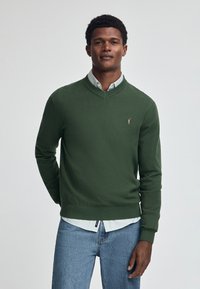 Grön v-ringad tröja i stickad kvalitet med ribbad nederkant och ärmslut. I kombination med en ljusblå skjorta med krage under.