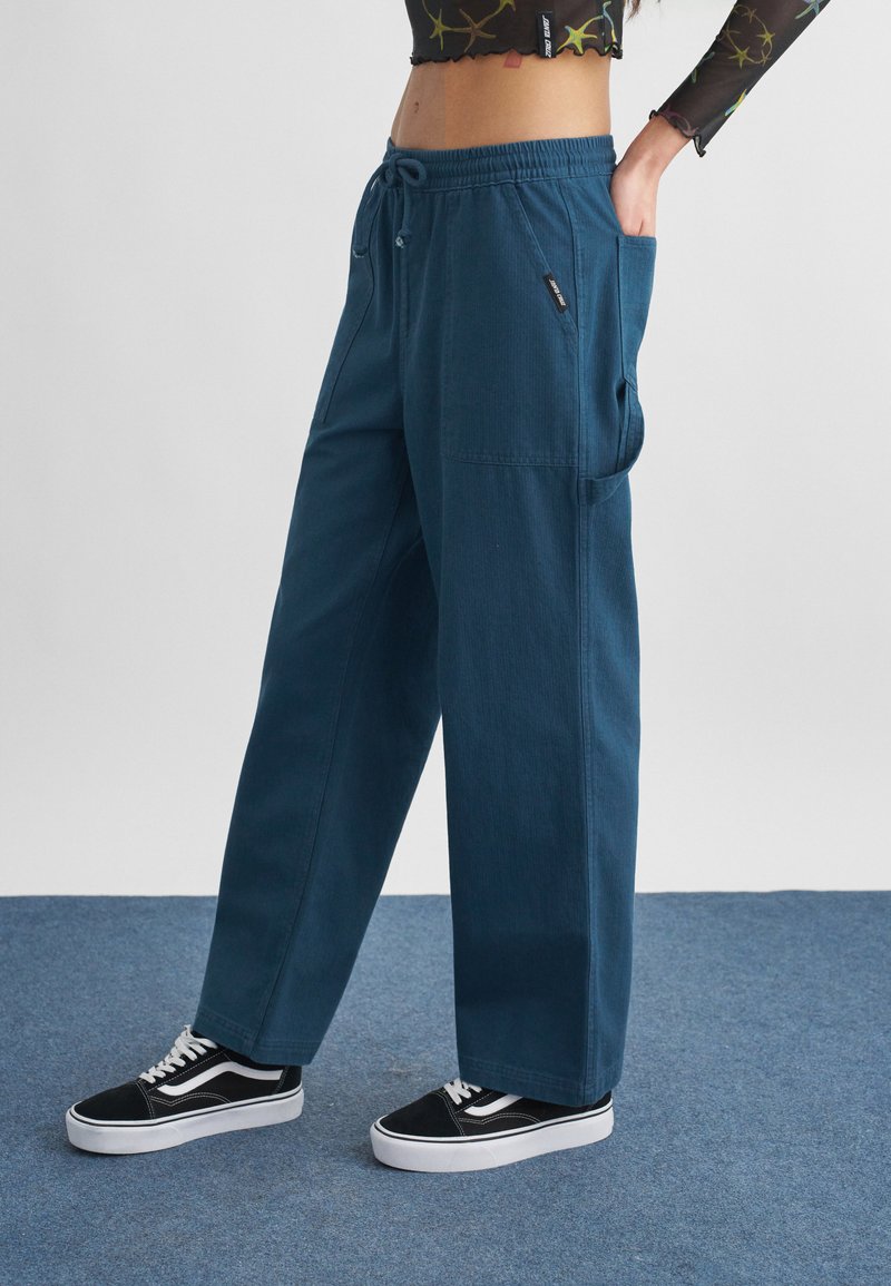 Άτομο που φοράει φαρδιά μπλε παντελόνια με ελαστική μέση και πλαϊνές τσέπες, συνδυασμένα με μαύρα και λευκά αθλητικά παπούτσια, στέκεται σε μπλε χαλί.