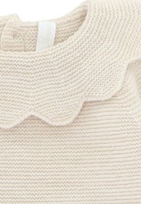 Suéter de punto en suave color crema, con un cuello festoneado y una etiqueta. La textura es suave con sutiles estrías en la tela.