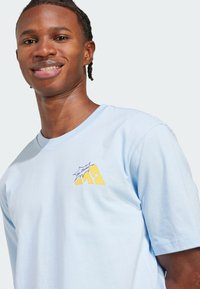 Světle modré bavlněné tričko s krátkými rukávy. Na levé části hrudi se nachází žlutý grafický logo, které obsahuje text a stylizovaný design.