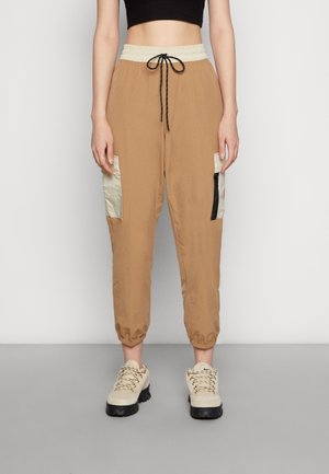 Personne portant un pantalon jogger cargo beige avec des poches beiges, un cordon noir, des baskets beiges épaisses et un top court noir, debout sur un fond blanc.
