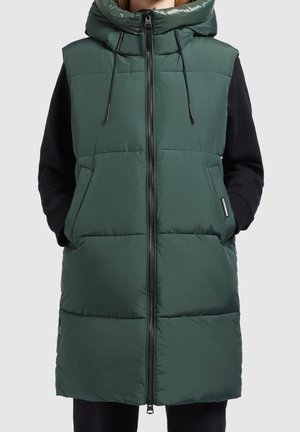 Frau trägt eine ärmellose, dunkelgrüne wattierte Weste mit Kapuze und schwarzen Ärmeln, Hände in den Taschen, Reißverschluss vorne geschlossen.