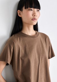 Camiseta de algodón marrón con manga corta, escote redondo y textura suave, presentada contra un fondo neutro.