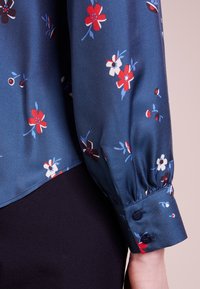Kwiatowa bluzka w ciemnoniebieskim satynie, z czerwonymi, białymi i niebieskimi kwiatami. Długie, marszczone rękawy z zapięciem na guziki. Gładka tekstura.