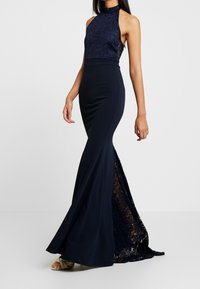 Kobieta w bezrękawowej granatowej sukni wieczorowej z koronkowym gorsetem i koronkowymi panelami, stojąca na białym tle.