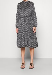 Robe à motifs noir et blanc avec des manches longues, un col haut et des volants en cascade au niveau de l'ourlet, associée à des sandales minimalistes noires.