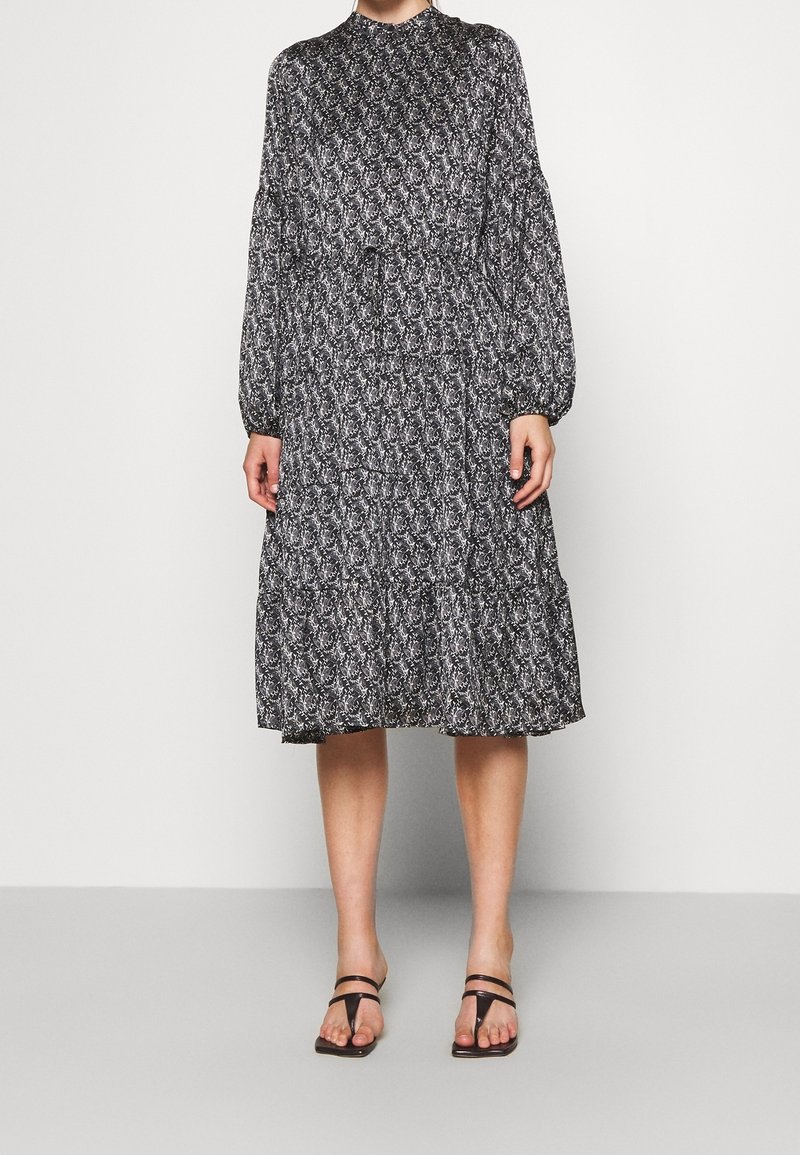 Robe à motifs noir et blanc avec des manches longues, un col haut et des volants en cascade au niveau de l'ourlet, associée à des sandales minimalistes noires.