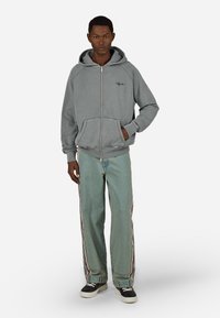 Pelēks zip-up hoody ar aukliņu kapuci un kangaroo kabatām, savienots ar gaismi zilām džinsiem ar kontrastējošām svītrām un baltām kurpēm.