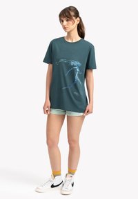 Teal katoenen t-shirt met korte mouwen, met een blauwe grafische ontwerp. Gecombineerd met lichtgroene shorts en witte hoge sneakers.