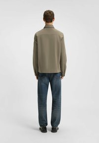 Oliven grøn skjorte med en lige skæring, lange ærmer og spidse krave, parret med løstsiddende blå jeans og sorte sko.