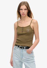 oliwkowy rybny top na ramiączkach z falbaniastym dekoltem, z przodu guziki i nadrukowanym tekstem "SUPERDRY 19 ATHL." W zestawie z jasnoniebieskimi dżinsami i czarnym paskiem.