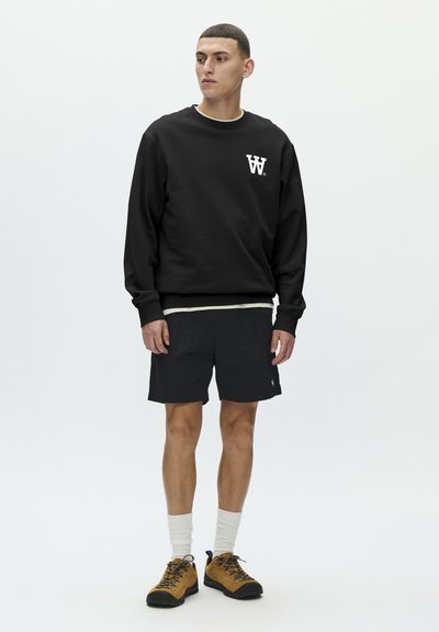 Sweat-shirt noir avec un logo blanc, associé à un short foncé. Le mannequin porte des chaussettes blanches et des chaussures noires et jaunes. Design simple et décontracté.