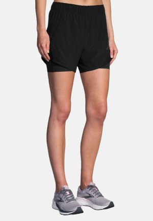 Persona vistiendo pantalones cortos deportivos negros y zapatillas de correr grises, de pie con las manos relajadas a los lados contra un fondo liso.