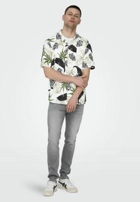 Camisa de manga corta blanca con patrones de hojas verdes y negras, combinada con jeans gris claro y zapatillas blancas con toques negros.