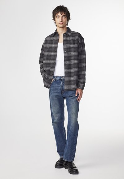 Sort og gråternet flannel skjorte over en hvid tanktop, parret med blå lige ben jeans og sorte sko.
