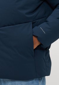 Hand in pocket of navy blue padded jacket with "JACK & JONES" logo on sleeve, worn over blue jeans.