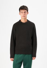 Maglione marrone scuro a coste con scollatura rotonda, maniche lunghe e vestibilità rilassata, abbinato a pantaloni verdi. Texture morbida e design sobrio.