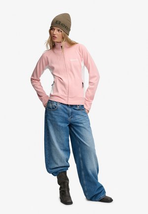 Kvinde iført en lyserød lynlåstrøje, løse blå jeans, sorte støvler og en brun beanie med "Bench" logo på en hvid baggrund.