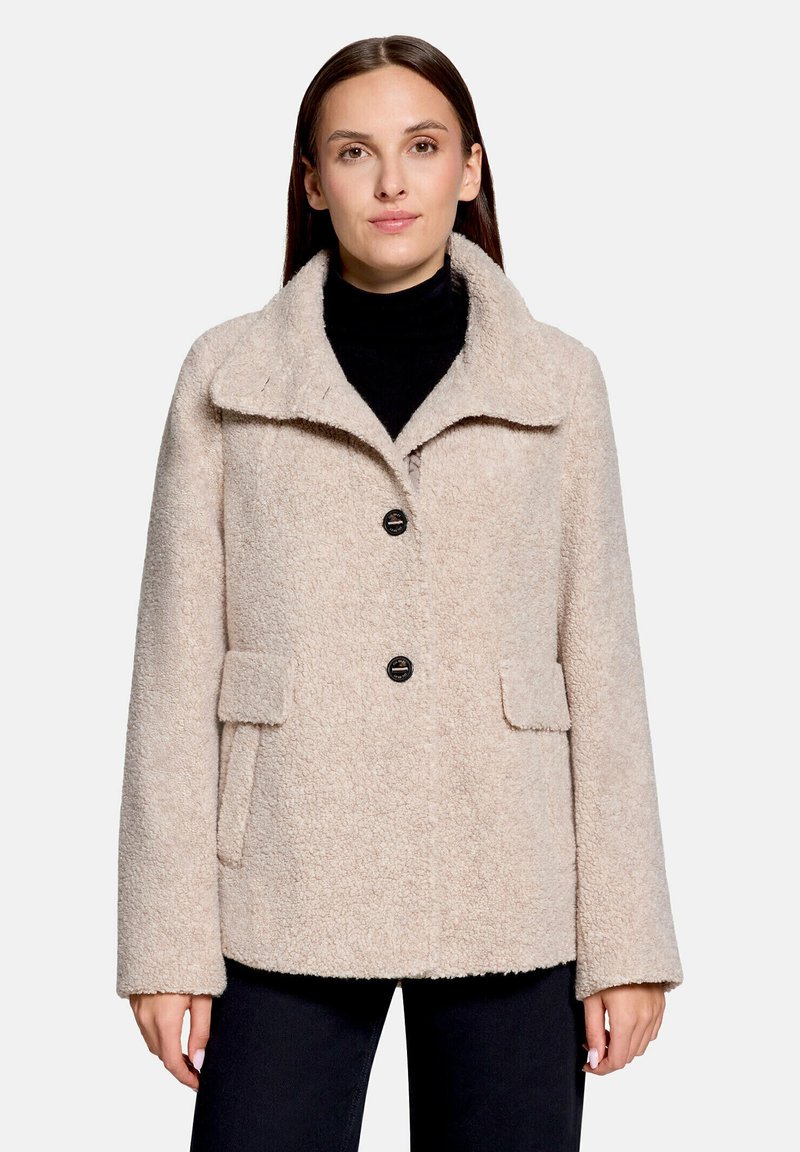 Femme portant un manteau en laine beige texturé avec un grand col et des boutons noirs sur un pull à col roulé noir.