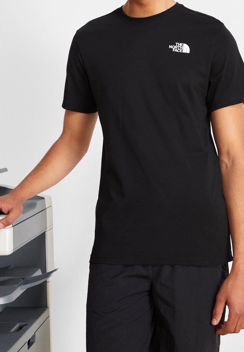 Black short-sleeve t-shirt made of cotton, featuring a white logo on the upper left chest. Worn with dark pants, standing next to a printer.