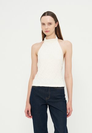 Jeune femme aux longs cheveux bruns portant un top dos-nu en dentelle blanche et un jean bleu foncé sur un fond blanc uni.