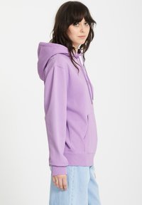 Luźny, lawendowy hoodie z kieszenią z przodu, kapturem ze sznurkiem i ściągaczami na rękawach. W połączeniu z jasnoniebieskimi dżinsami.