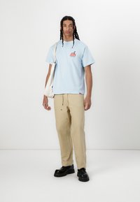Light blue short-sleeve t-shirt with graphic print, beige pants with pockets, black chunky shoes, and a white tote bag.