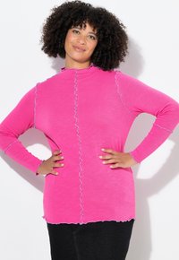 Pitkähihainen vaaleanpunainen paita, jossa on röyhelöidyt reunat ja kontrastiväriset siniset tikit, korkea kaulus ja hieman löysä istuvuus.