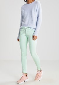 Pull bleu clair en tissu pelucheux, jean ajusté vert menthe avec fermetures éclair latérales, et baskets roses à enfiler avec semelles blanches.